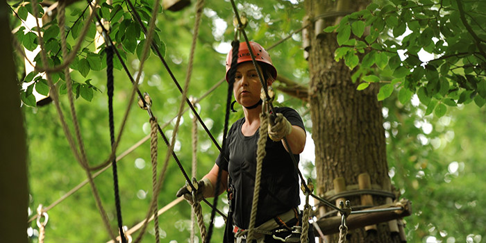 Une personne équipée d'un casque rouge s'aventure sur un parcours d'accrobranche, surmontant des obstacles dans une forêt luxuriante.