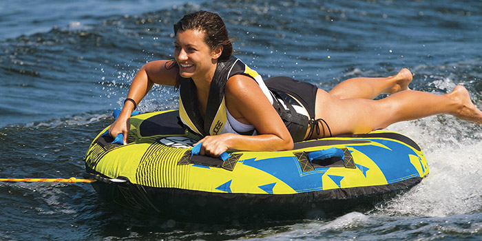 Femme souriante sur bouée tractée en été sur un lac, éclaboussures et aventure aquatique sous le soleil.