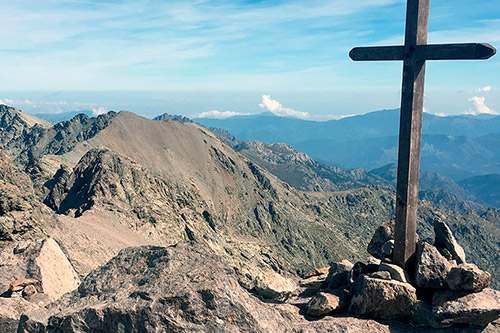 "Croix en bois au sommet d'une montagne avec un paysage panoramique, ciel bleu et crêtes rocheuses."