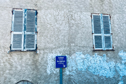 Image de la façade d'un bâtiment historique à Cargèse avec volets bleus usés et panneau indicatif « le Port », illustrant le charme du village corse et sa proximité avec la mer.