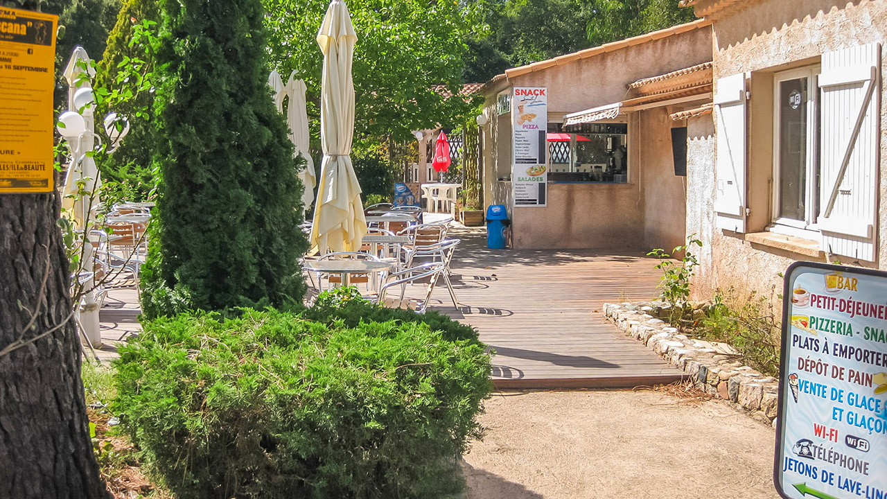 Terrasse d'un snack ombragée par de grands arbres, idéale pour se détendre avec un café ou une pizza en pleine nature, créant une ambiance estivale et conviviale.