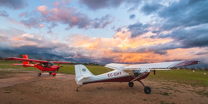 Deux avions légers éclairés par un coucher de soleil doré sur un terrain d'aviation, entourés de nuages bleus et oranges, évoquent l'aventure aérienne.