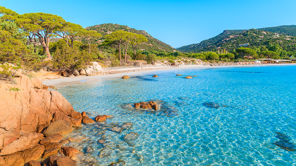 Paysage côtier serein avec plage, mer bleu azur et collines verdoyantes en arrière-plan.