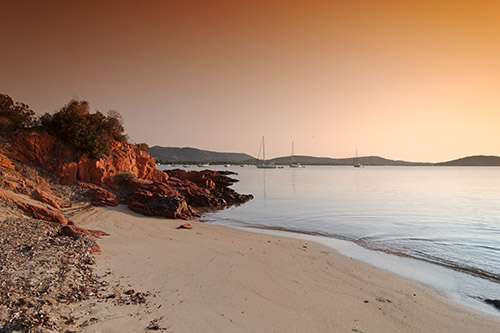 Coucher de soleil enchanteur sur une plage calme avec voiliers à l'horizon, reflet doré sur l'eau.