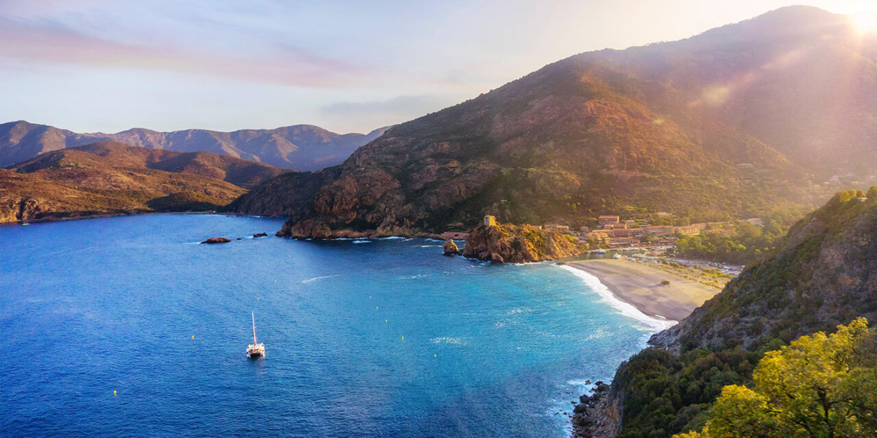 Paysage de Corse au crépuscule : montagnes majestueuses et mer azur illuminées par un coucher de soleil doré, invitant à l'évasion naturelle.