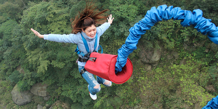 Femme souriant pendant un saut élastique au-dessus d'une forêt verdoyante, symbolisant joie, adrénaline et liberté.