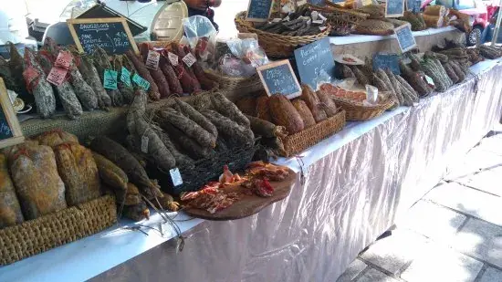 charcuterie-corse-aop-porc-noir Stand de marché en plein air présentant une grande variété de charcuterie corse traditionnelle avec des saucissons artisanaux suspendus et des pièces de salaison exposées dans des paniers en osier.