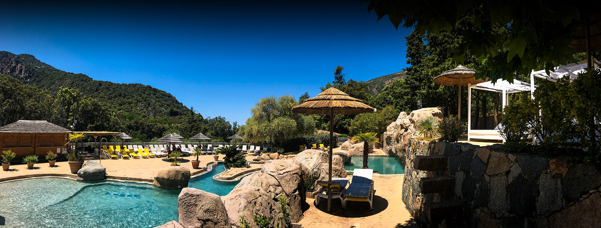 Piscine cristalline entourée de verdure et de montagnes avec des chaises longues et parasols en paille sous le soleil, créant une ambiance détente et idyllique.