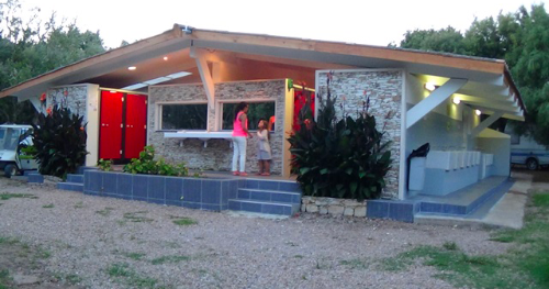 Bâtiment de camping moderne avec murs en pierre et portes rouges illuminées au crépuscule, entouré de plantes luxuriantes, créant une atmosphère chaleureuse et conviviale pour les campeurs.