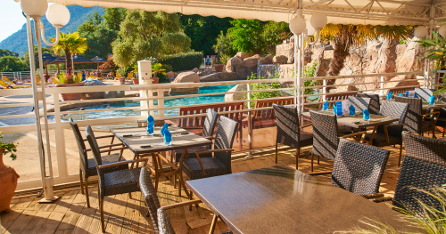 Terrasse de restaurant avec vue sur une piscine étincelante entourée de verdure et de palmiers, parfaite pour un repas convivial en extérieur.