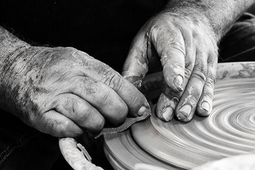 Image en noir et blanc illustrant un artisan potier concentré, ses mains ridées formant délicatement l'argile sur un tour de potier. Chaque geste révèle une expertise et une passion pour cet art traditionnel.