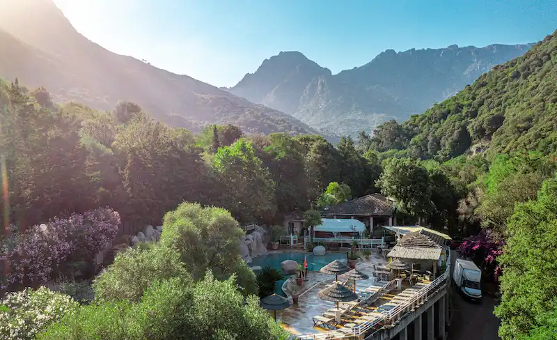 Terrasse illuminée du bar-restaurant du camping surplombant la piscine avec les montagnes en arrière-plan.