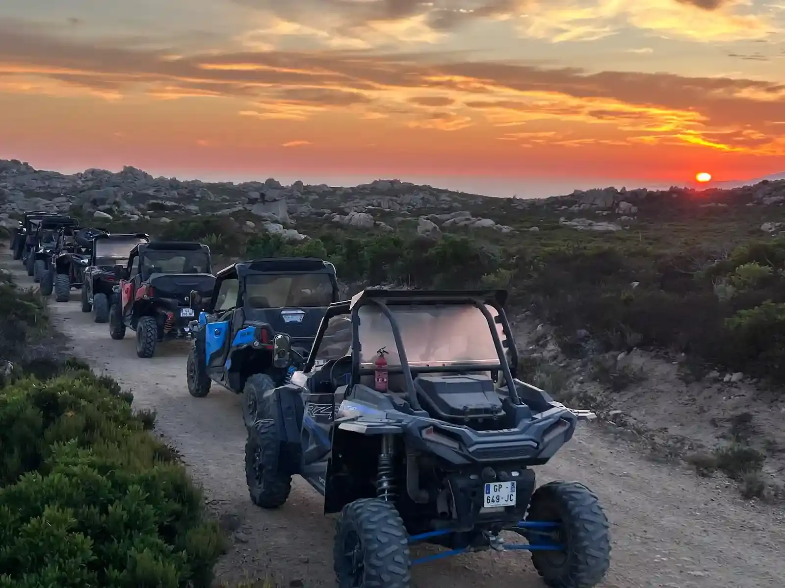 File indienne de buggies et quads tout-terrain sur un sentier du maquis corse face à un coucher de soleil orange flamboyant.