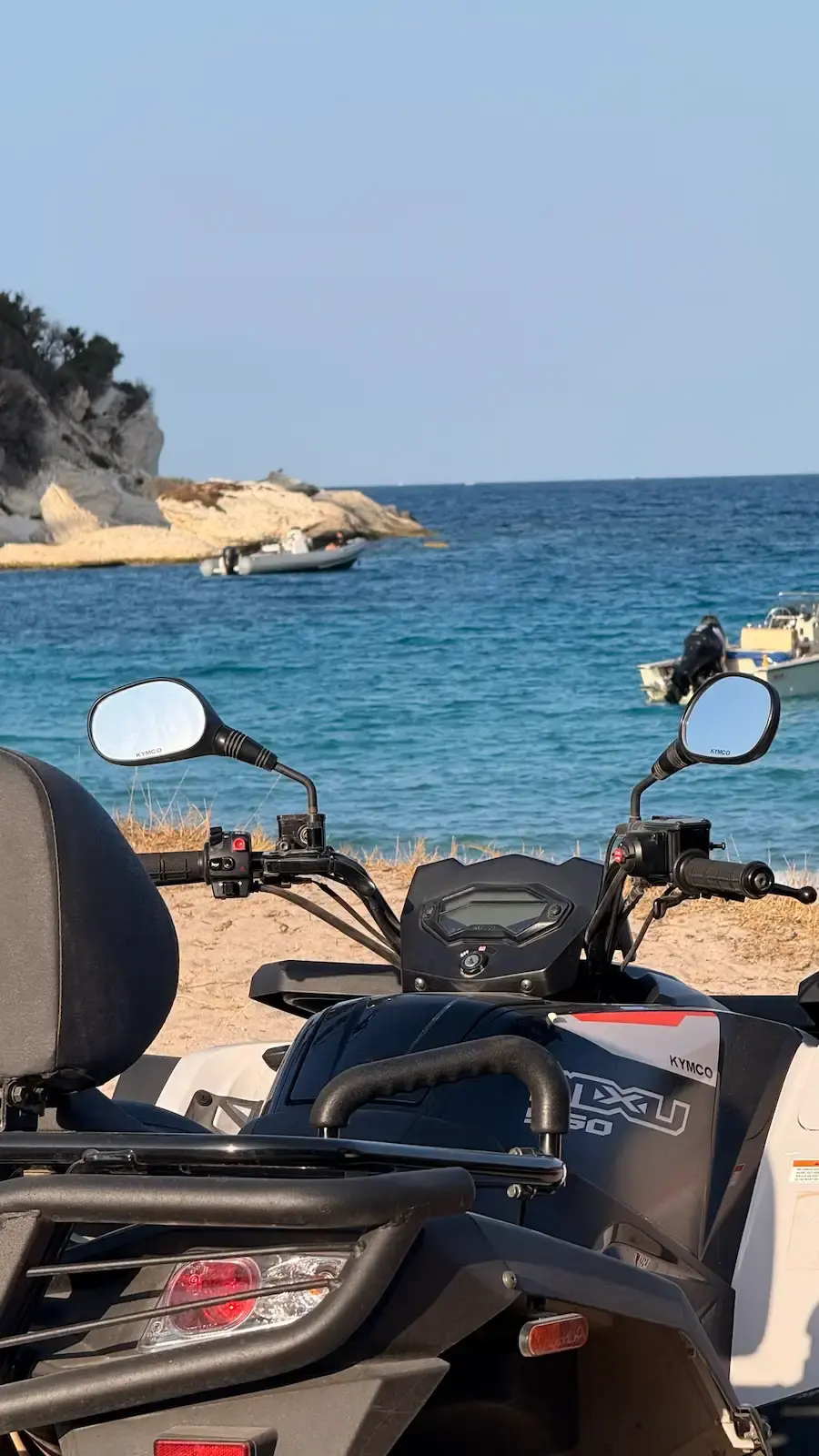 Gros plan sur le tableau de bord d'un quad Kymco MXU 550 stationné sur le sable face à la mer turquoise et des bateaux.