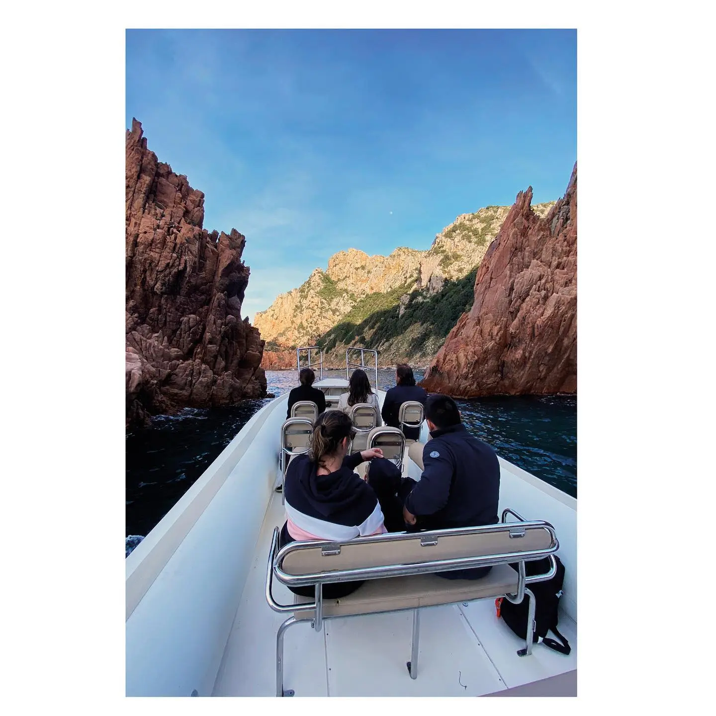 Vue depuis l'arrière du bateau d'excursion "Le Goéland" montrant plusieurs passagers installés sur des sièges, naviguant sur une mer bleue entre d'imposantes falaises rocheuses rouges de la réserve de Scandola en Corse, sous un ciel bleu.