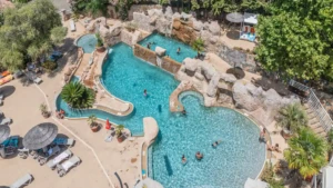 Vue aérienne du grand complexe aquatique de type lagon au Camping Les Oliviers à Porto, en Corse-du-Sud. On y voit des baigneurs dans les différentes piscines reliées par des cascades artificielles en rochers, des transats et parasols en paillote sur la terrasse ensoleillée, entourés d'une végétation méditerranéenne luxuriante.