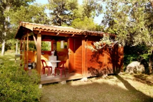 Un chalet en bois avec toit en tuiles au Camping U Stabiacciu, Porto-Vecchio. Il dispose d'une terrasse couverte avec deux chaises rouges et une table blanche, entouré de verdure et de maquis corse.