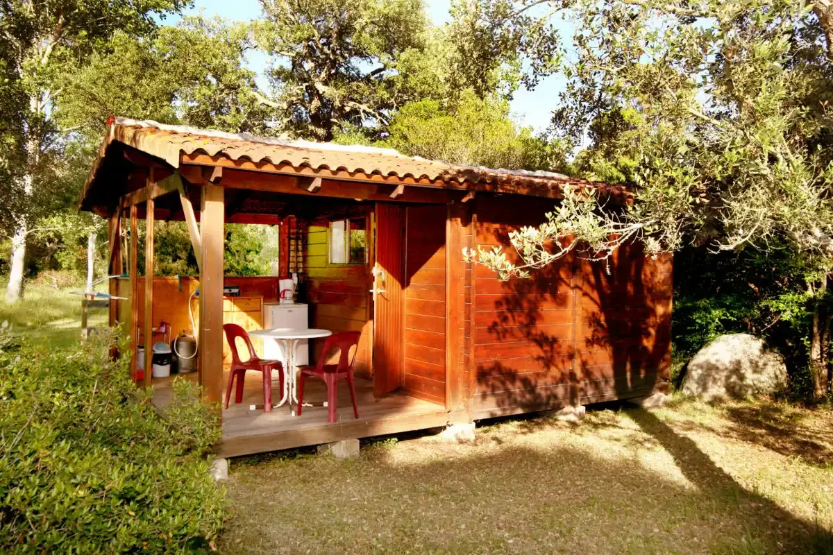 Un chalet en bois avec toit en tuiles au Camping U Stabiacciu, Porto-Vecchio. Il dispose d'une terrasse couverte avec deux chaises rouges et une table blanche, entouré de verdure et de maquis corse.
