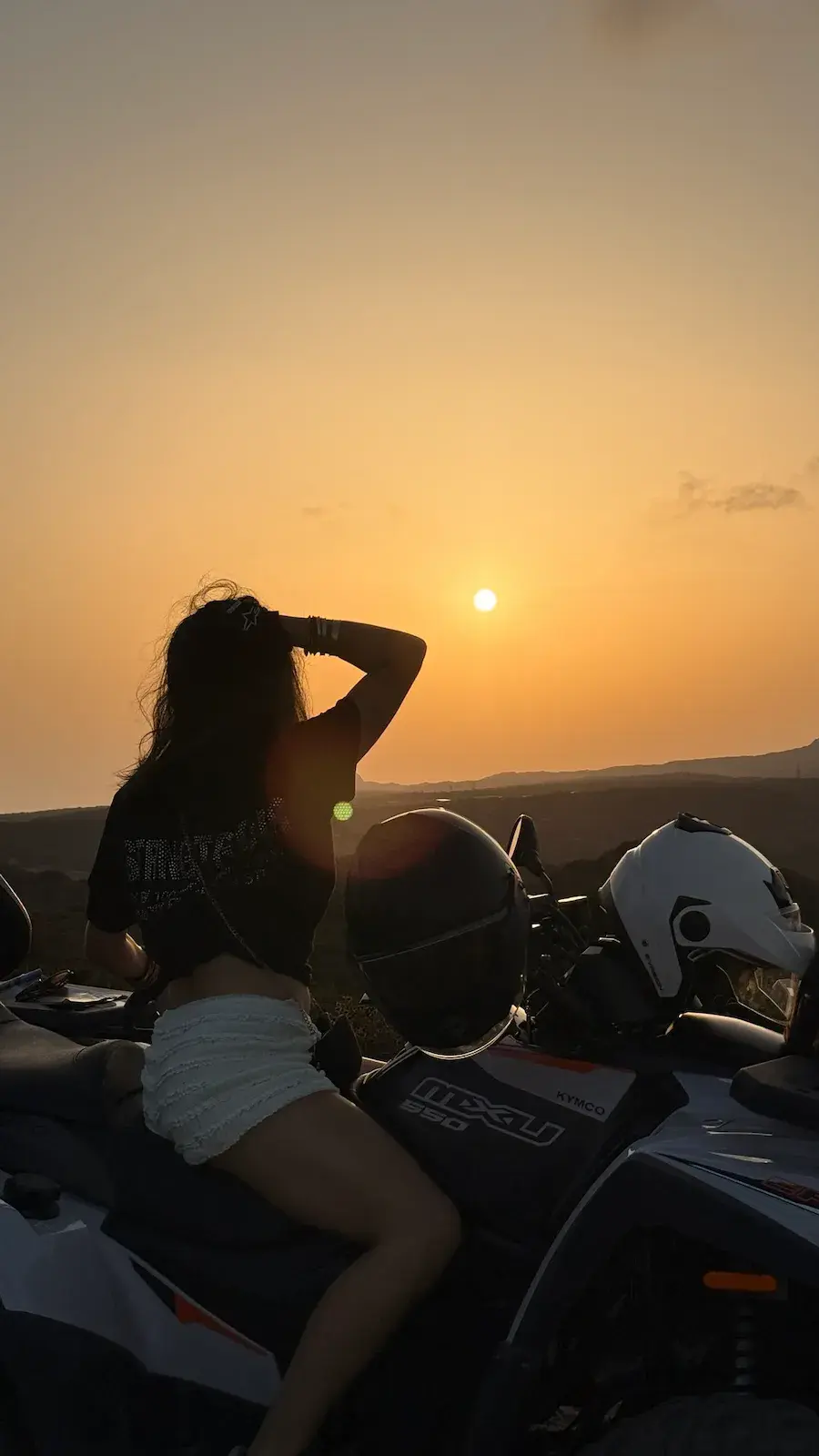 Femme assise sur un quad Kymco MXU 550 admirant le coucher de soleil à l'horizon, casques de protection posés sur le véhicule.