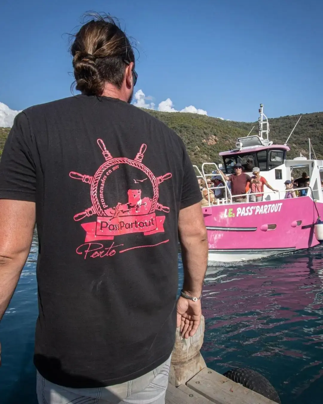 Vue de dos d'un capitaine portant un t-shirt noir logoté Le Pass'Partout Porto, regardant son bateau s'approcher du quai.