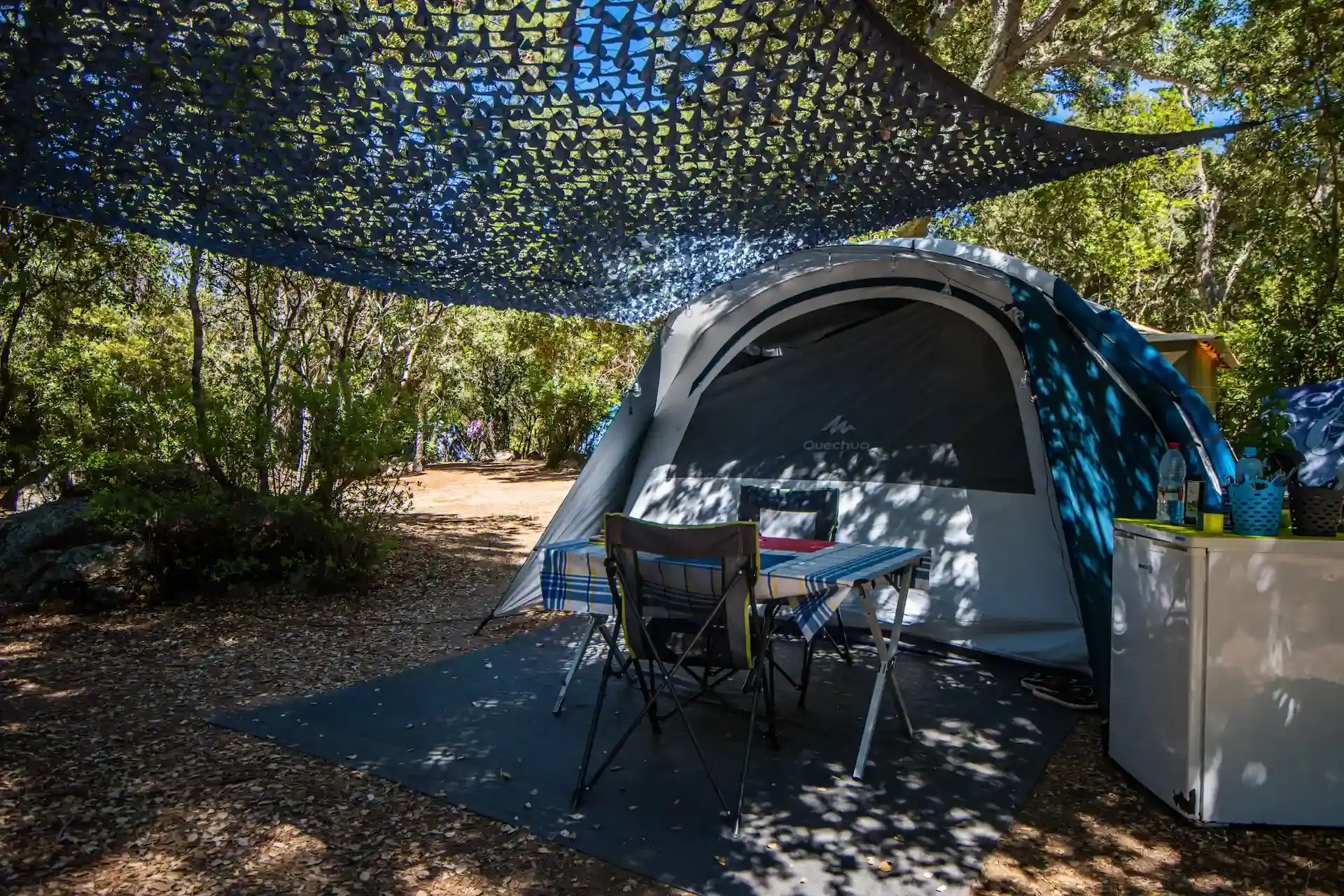 Emplacement de camping ombragé avec grande tente Quechua installée, table de jardin et petit réfrigérateur.