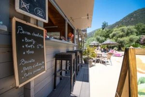 Le bar du Camping Les Oliviers à Porto vu depuis la terrasse en deck. Un grand tableau noir suspendu affiche le menu des collations en français ("Barres chocolatées" et "Biscuits"), à côté de tabourets de bar en résine tressée. En arrière-plan, la terrasse du restaurant avec des tables, des chaises et des parasols en paillote donne sur la piscine et les montagnes environnantes.