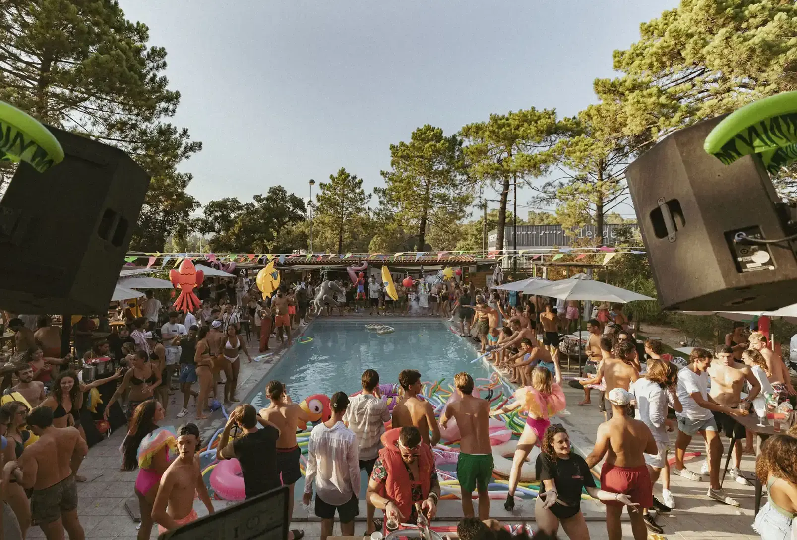 Une foule de personnes autour de la piscine du Camping U Stabiacciu à Porto-Vecchio lors d'une pool party. Des haut-parleurs noirs flanquent la scène avec un DJ au fond sous une structure, sous un ciel dégagé.