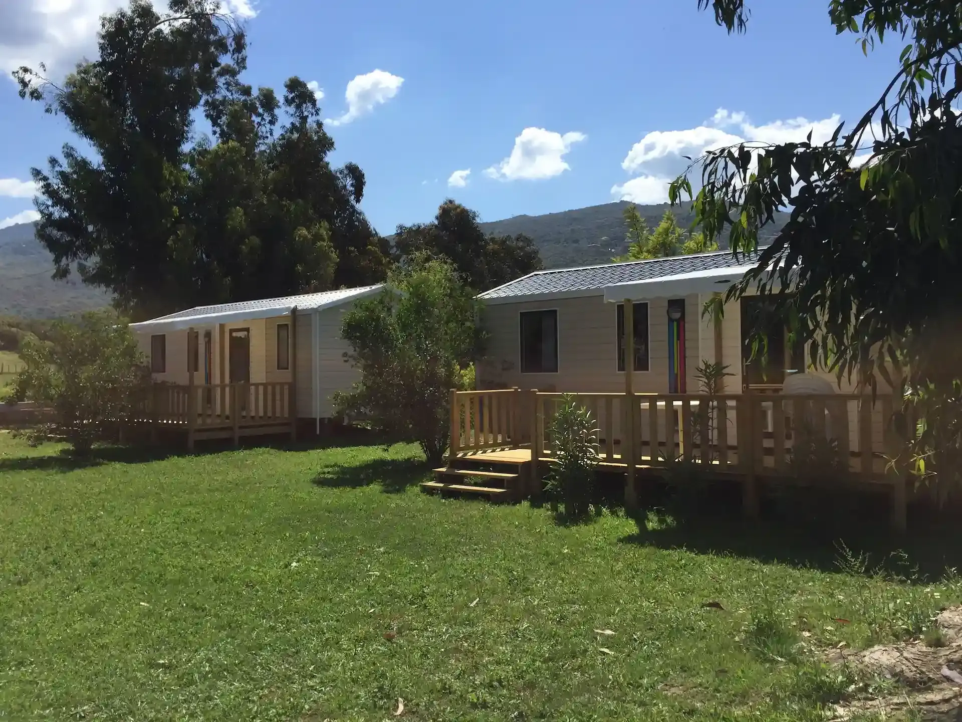 Deux mobile-homes avec terrasses en bois sur une grande pelouse verte au Camping Milella à Propriano, avec des montagnes et un ciel nuageux en arrière-plan.