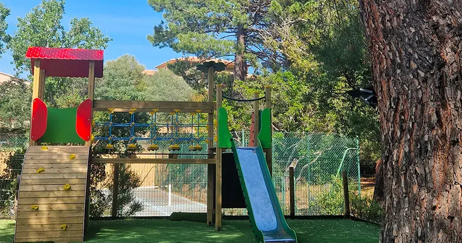 Aire de jeux extérieure sécurisée pour enfants au Camping U Pinarellu à Porto-Vecchio, comprenant un toboggan bleu, un pont de cordes et une structure d'escalade en bois sur gazon artificiel.