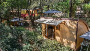 Un chalet en bois avec une terrasse couverte au Camping Les Oliviers à Porto, niché sur une colline arborée face aux montagnes de Corse. En arrière-plan, on distingue une roulotte rouge et une autre zone de camping avec des parasols. Le chalet est entouré d'une végétation dense méditerranéenne, garantissant calme et intimité.