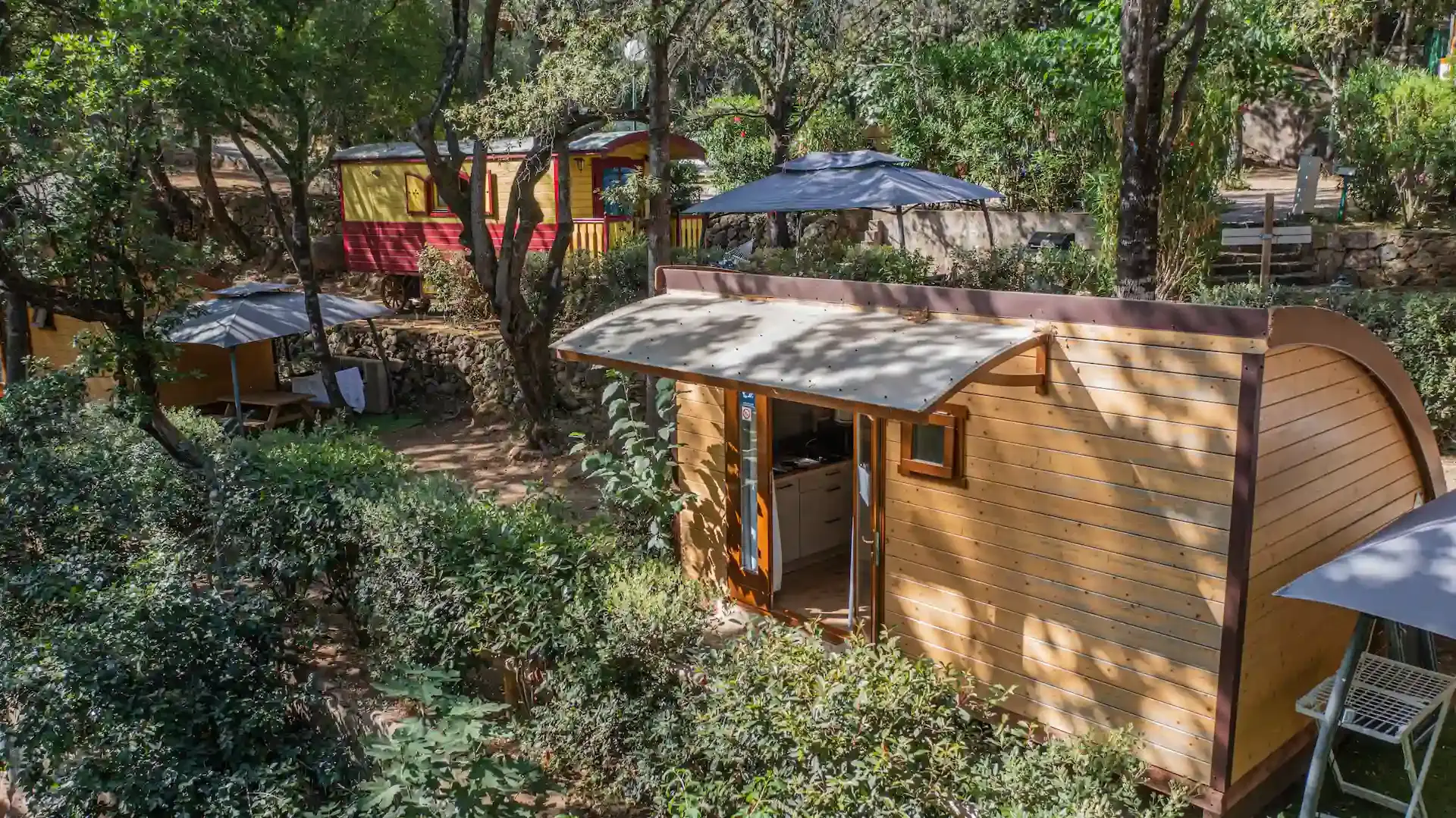 Un chalet en bois avec une terrasse couverte au Camping Les Oliviers à Porto, niché sur une colline arborée face aux montagnes de Corse. En arrière-plan, on distingue une roulotte rouge et une autre zone de camping avec des parasols. Le chalet est entouré d'une végétation dense méditerranéenne, garantissant calme et intimité.