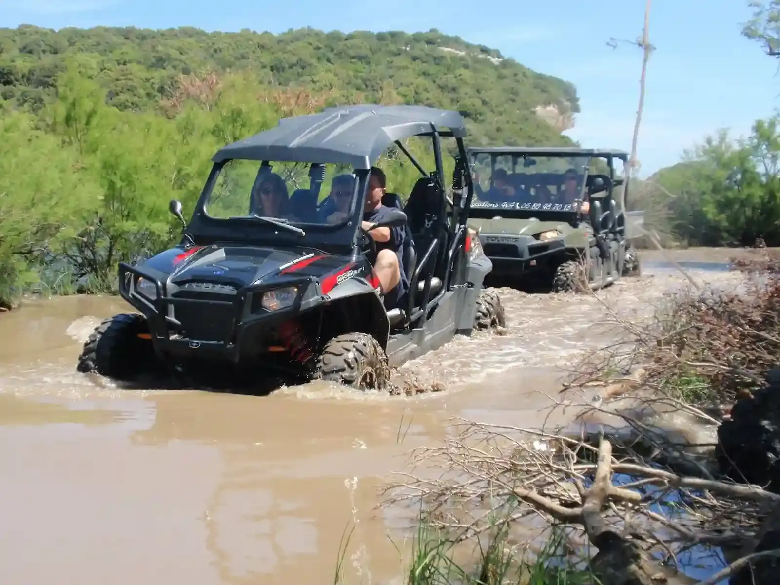 Deux buggies Polaris RZR franchissant un passage d'eau boueux dans une zone boisée sous un ciel dégagé.