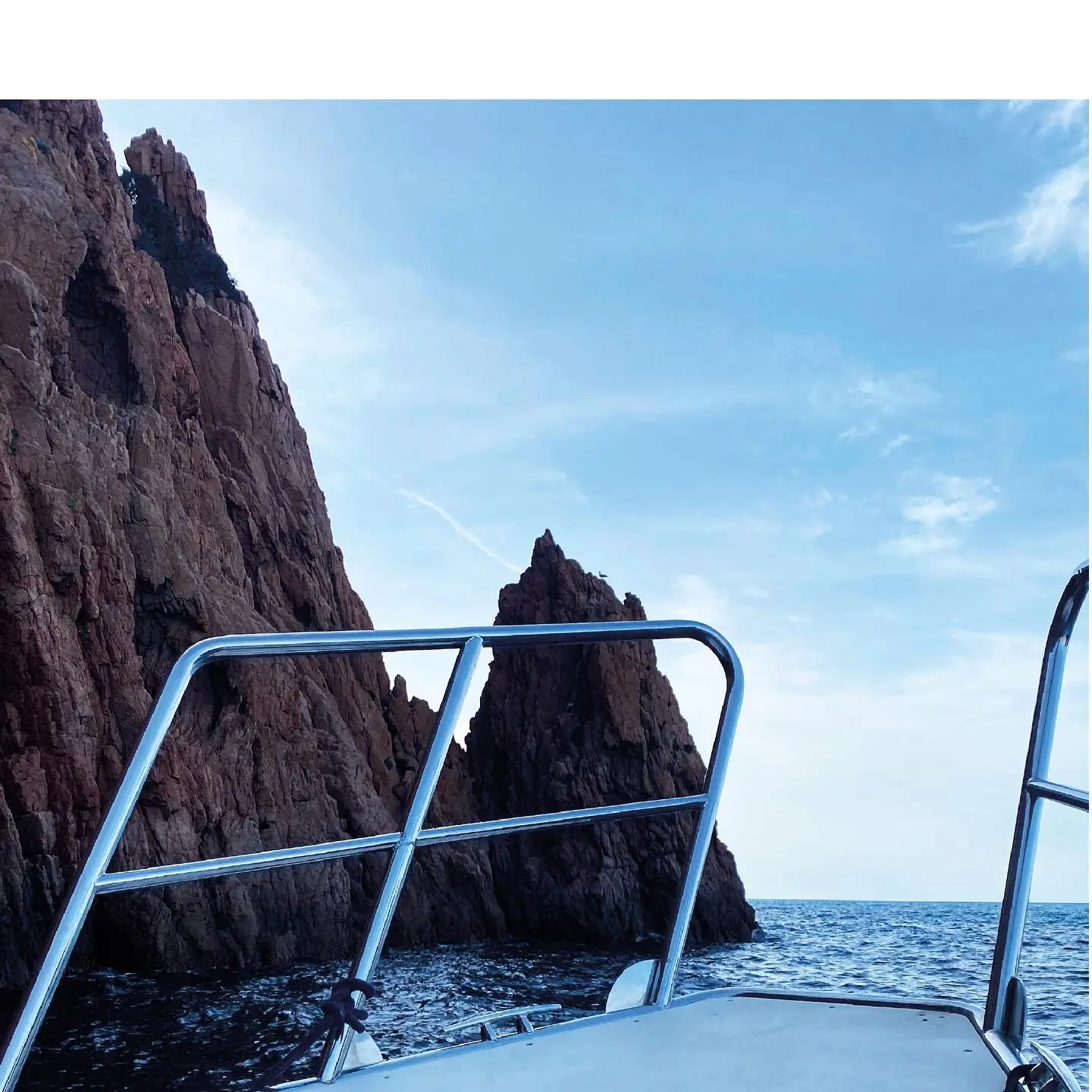 Vue depuis l'avant d'un bateau avec des rambardes en acier inoxydable, s'approchant d'impressionnantes falaises côtières rocheuses de couleur rouge et brune, sous un ciel bleu clair parsemé de quelques nuages, dans la réserve de Scandola en Corse.