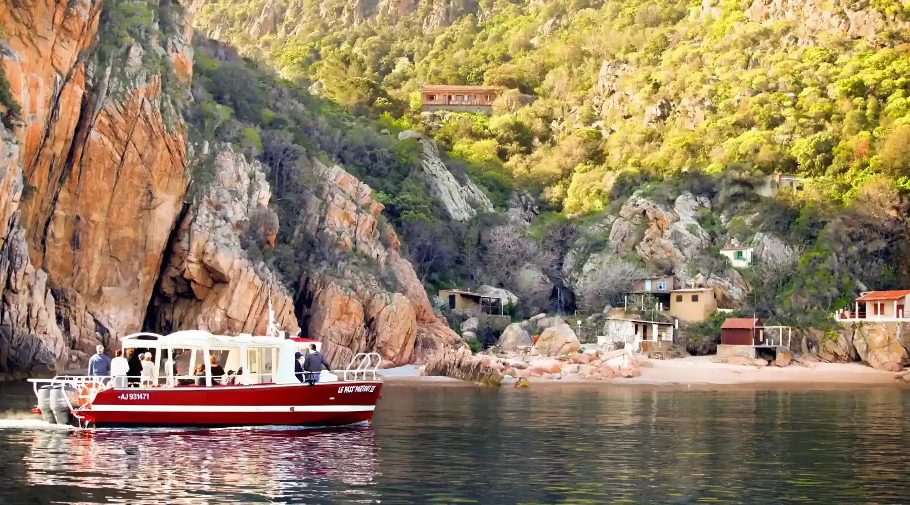 Bateau de promenade rouge et blanc naviguant devant une plage isolée et des maisons de pêcheurs au pied des montagnes corses.