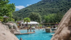 Vue d'ensemble de la piscine lagon du Camping Les Oliviers à Porto, Corse-du-Sud, prise depuis une terrasse surélevée. En arrière-plan, le restaurant du camping sous une tonnelle blanche, entouré de végétation et de parasols en paillote. Des baigneurs profitent de la piscine. Au-delà du camping, la vallée de Porto est entourée de montagnes couvertes de maquis, sous un ciel bleu clair.