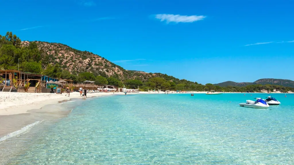 Vue de la plage de Santa Giulia près de Porto-Vecchio en Corse, avec son eau turquoise peu profonde, son sable fin et deux jet-skis amarrés, sous un ciel bleu pur.