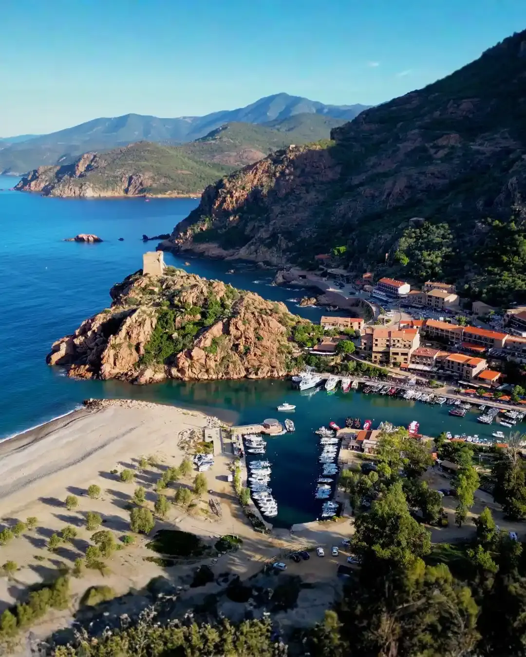 Vue aérienne panoramique de la marine de Porto en Corse, montrant le port de plaisance, la plage de galets et la tour génoise historique surplombant les eaux turquoise du golfe.