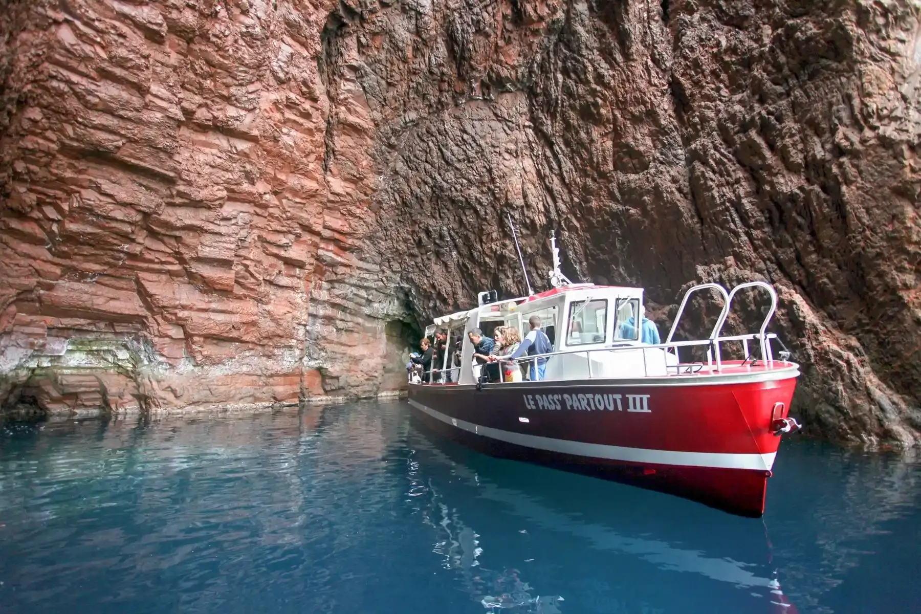 Le bateau Le Pass'Partout III pénétrant à l'intérieur d'une grotte marine aux parois de granit rouge dans les Calanques de Piana.