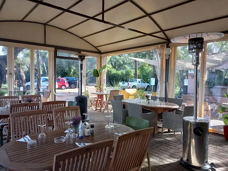 L'intérieur du restaurant du Camping U Stabiacciu sous une grande structure de terrasse couverte. Des tables en bois sont dressées avec verres et serviettes, entourées de chaises en rotin et de plantes. Des chauffages extérieurs sont visibles.