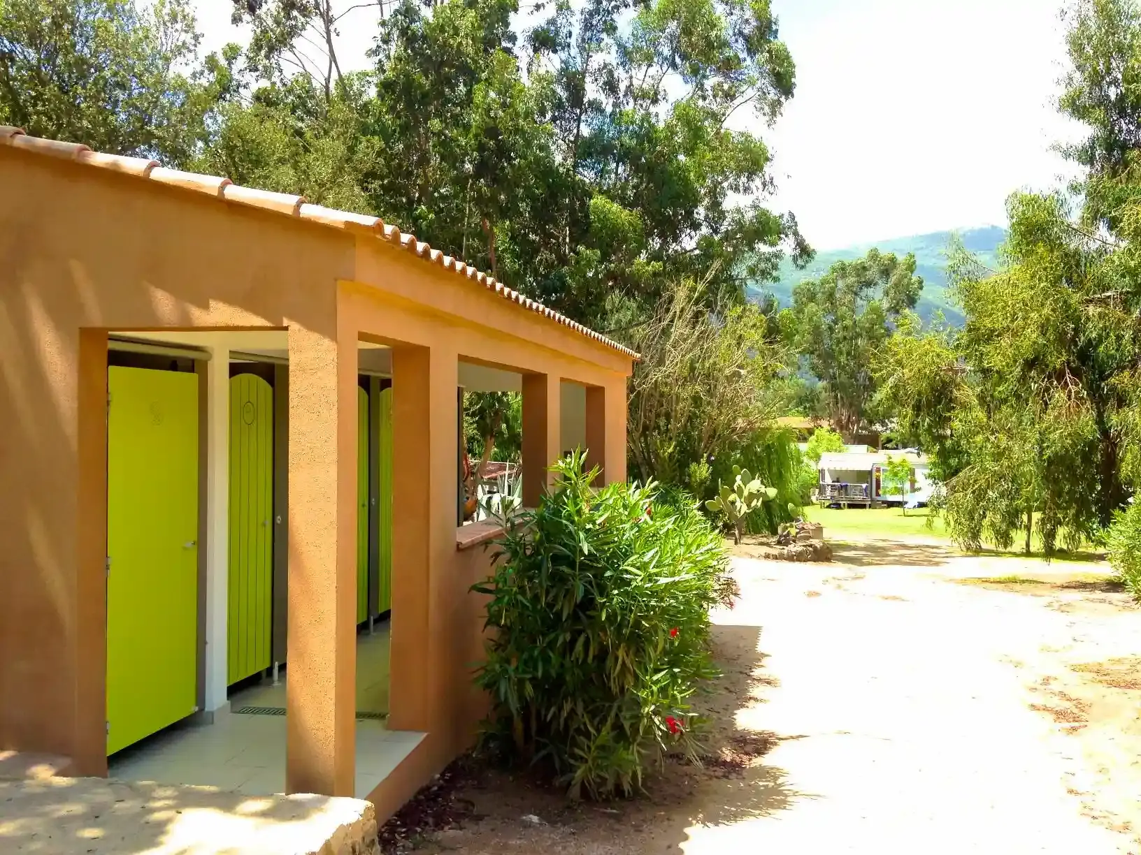 Vue d'un bloc sanitaire extérieur au Camping Milella à Propriano, Corse-du-Sud, avec des portes vert vif pour les douches et un laurier-rose en fleurs, sous un soleil éclatant.