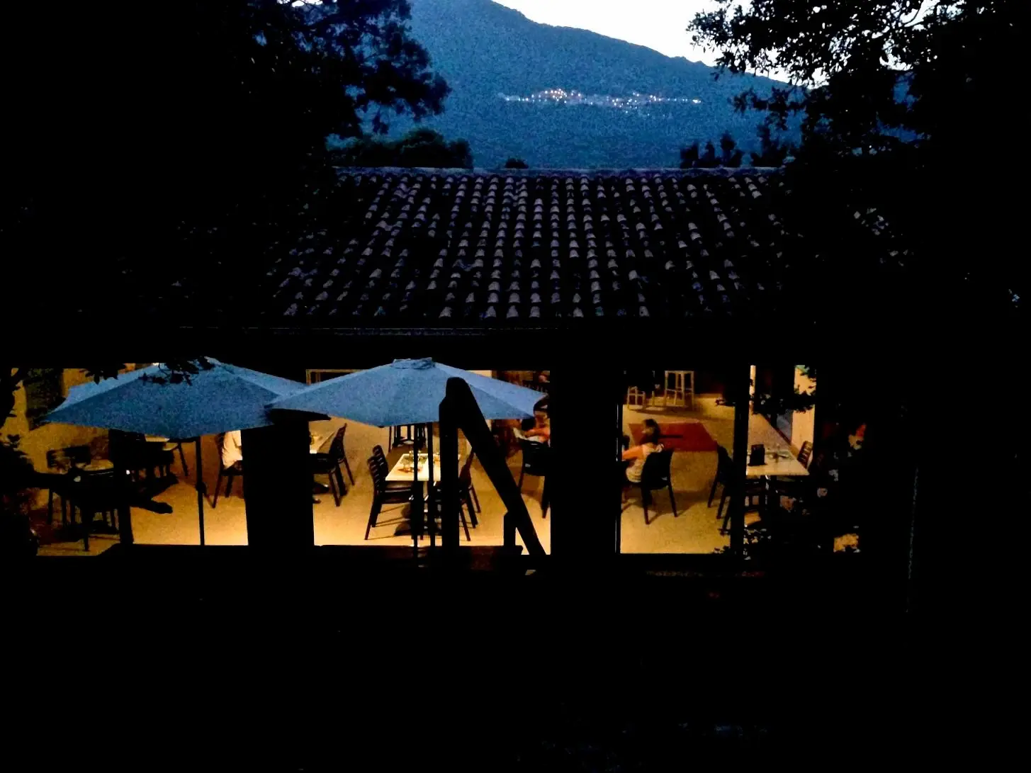 Terrasse du restaurant du Camping Milella à Propriano, Corse-du-Sud, en soirée. On distingue des tables dressées, des parasols bleus et des lumières chaudes à l'intérieur, avec une montagne sombre à l'horizon.