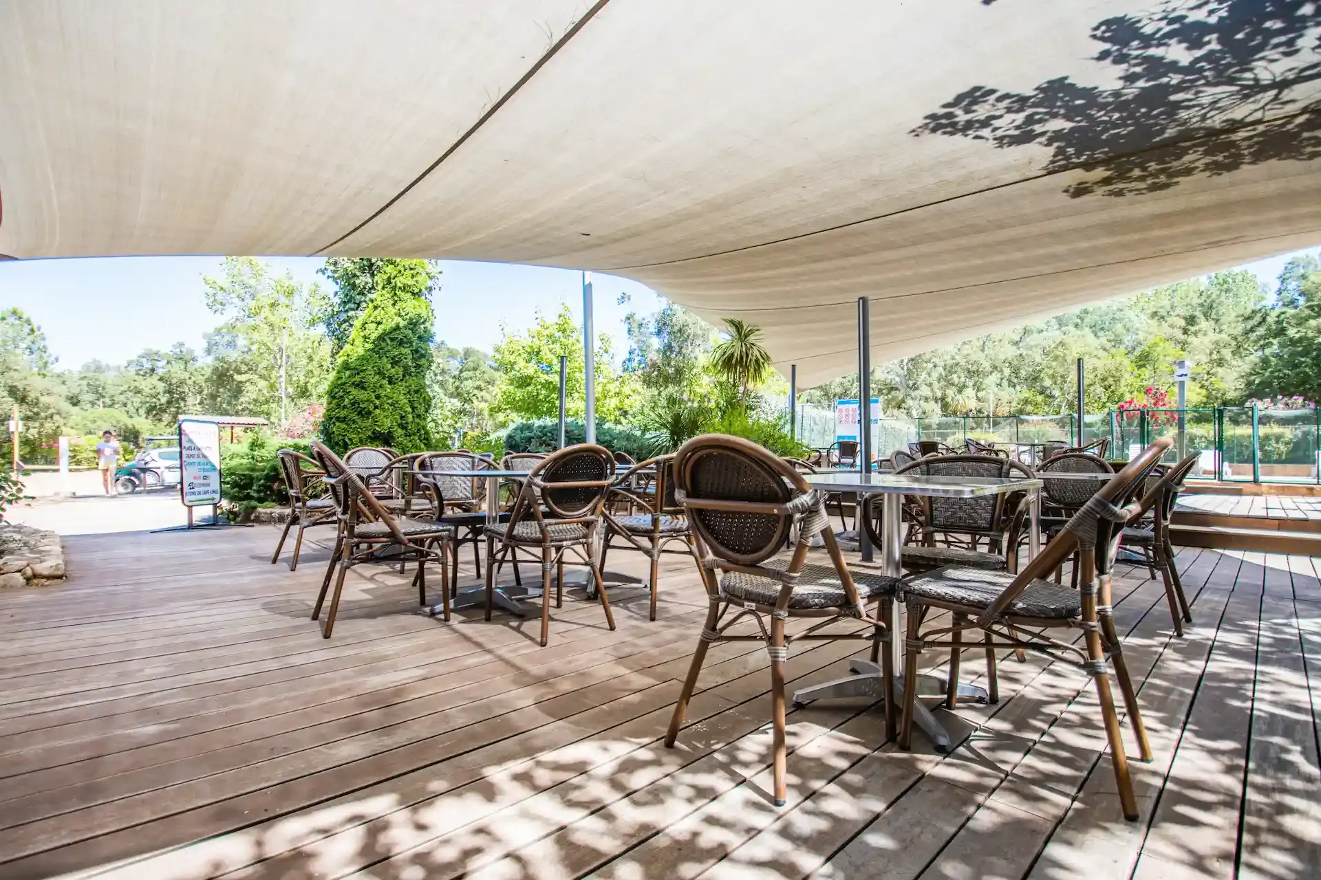 Terrasse de restaurant en bois sous une voile d'ombrage blanche avec vue sur la piscine et les jardins.