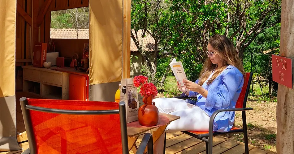 ne femme lisant un livre sur la terrasse en bois d'un lodge au Camping U Pinarellu, assise sur une chaise rouge devant une cuisine extérieure bien équipée et des plantes vertes.