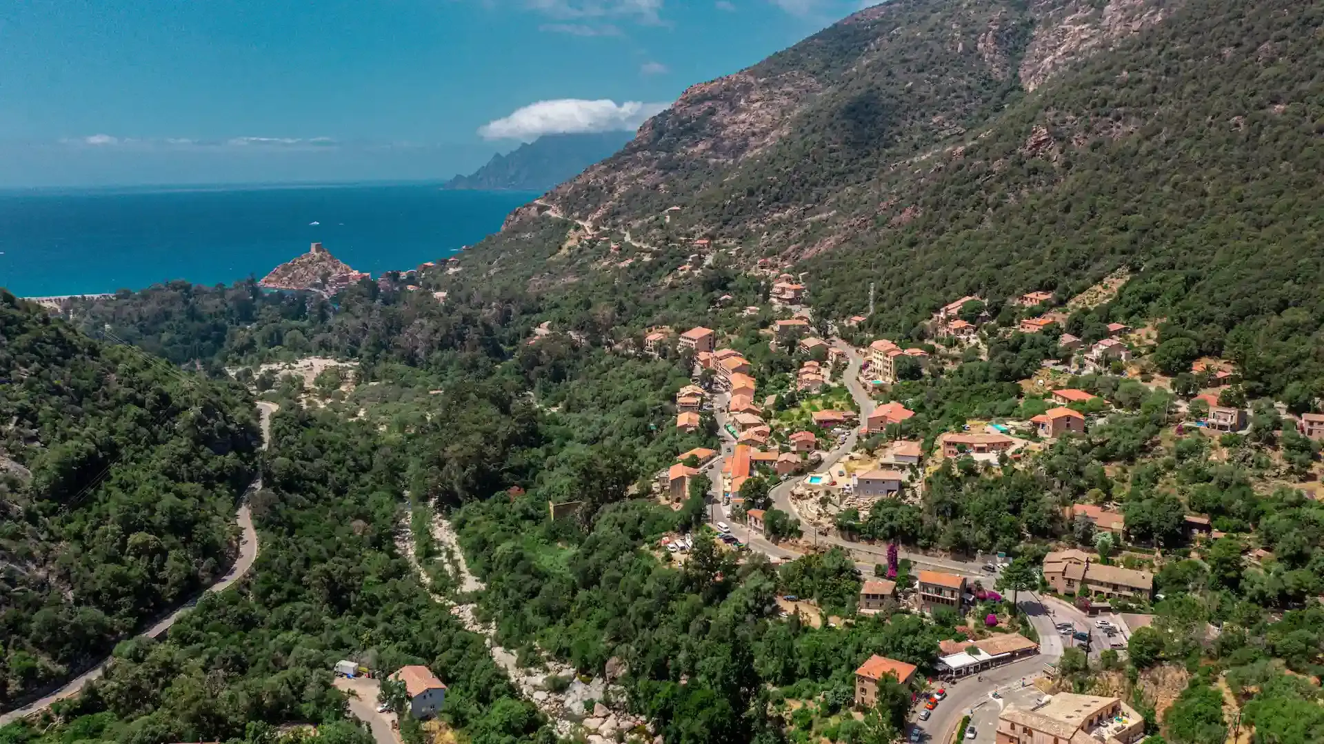 ue aérienne par drone du village de Porto et de son golfe en Corse-du-Sud. Le village aux toits rouges est niché dans une vallée verdoyante au pied de montagnes couvertes de maquis. La rivière Porto serpente vers la mer Méditerranée, où l'on distingue la tour génoise et la marina avec de nombreux bateaux de plaisance amarrés. Le Camping Les Oliviers est situé sur les hauteurs du village.