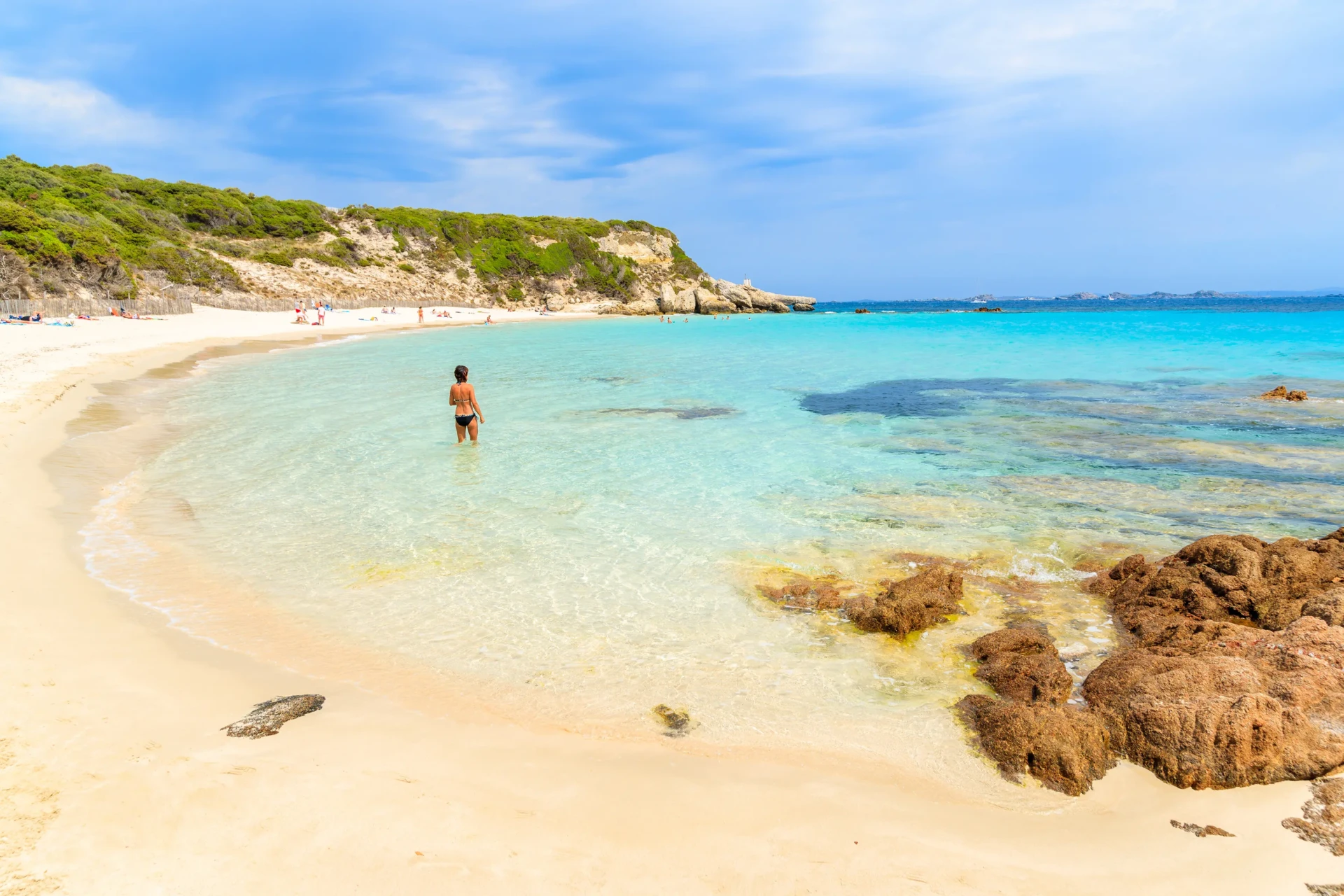 Silhouette d'une femme marchant seule dans l'eau turquoise et calme d'une crique de la plage du Grand Sperone, bordée de rochers au premier plan.