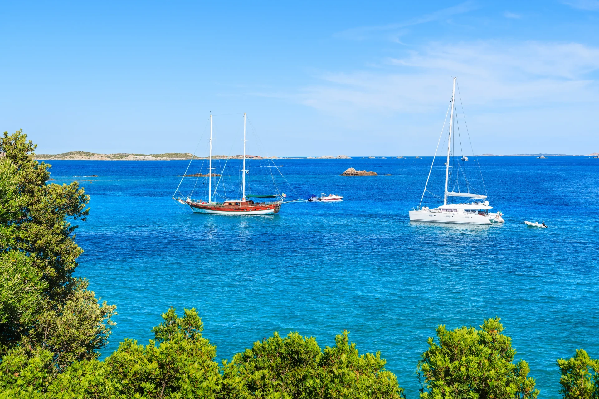 Voiliers et catamarans au mouillage dans les eaux bleu turquoise de la baie de Sperone à Bonifacio, photographiés à travers le maquis corse.