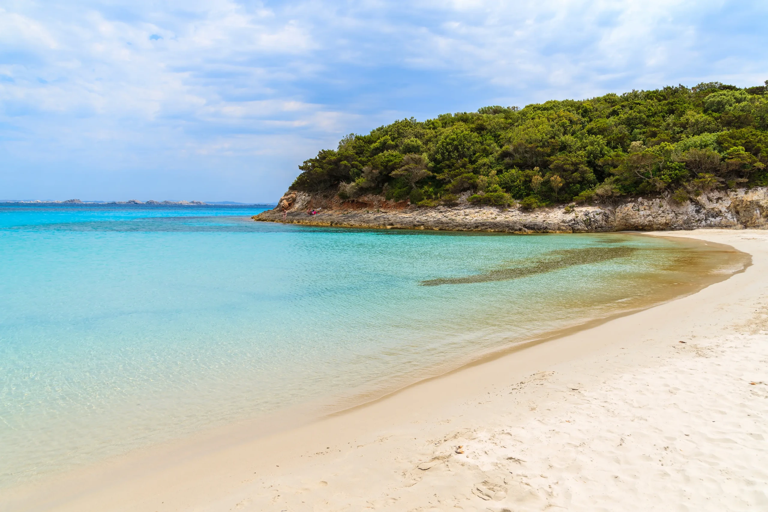 Plage de sable blanc déserte curvée vers une colline de maquis dense et une mer calme turquoise sous un ciel bleu parsemé de nuages blancs.