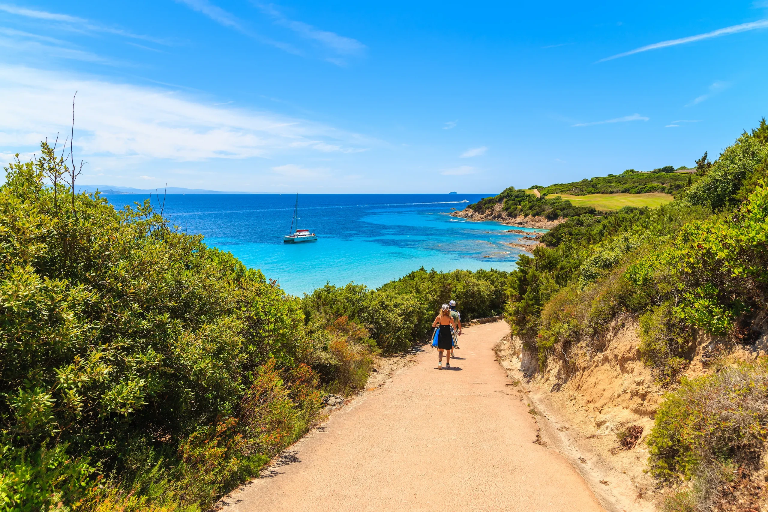 Deux randonneurs marchant sur le sentier côtier en terre battue qui descend vers la plage du Grand Sperone et sa mer turquoise.